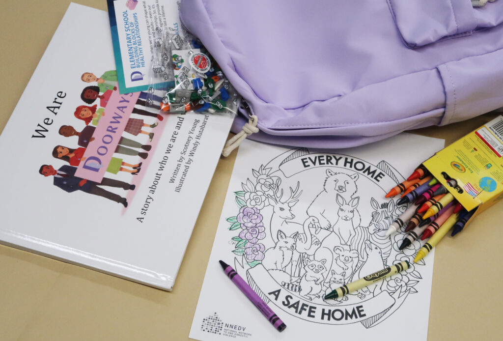 Light purple backpack with the children's book titled We Are Doorways, a coloring sheet that reads "every home a safe home," crayons, and a handout about the building blocks of healthy relationships, with a small set of building bricks