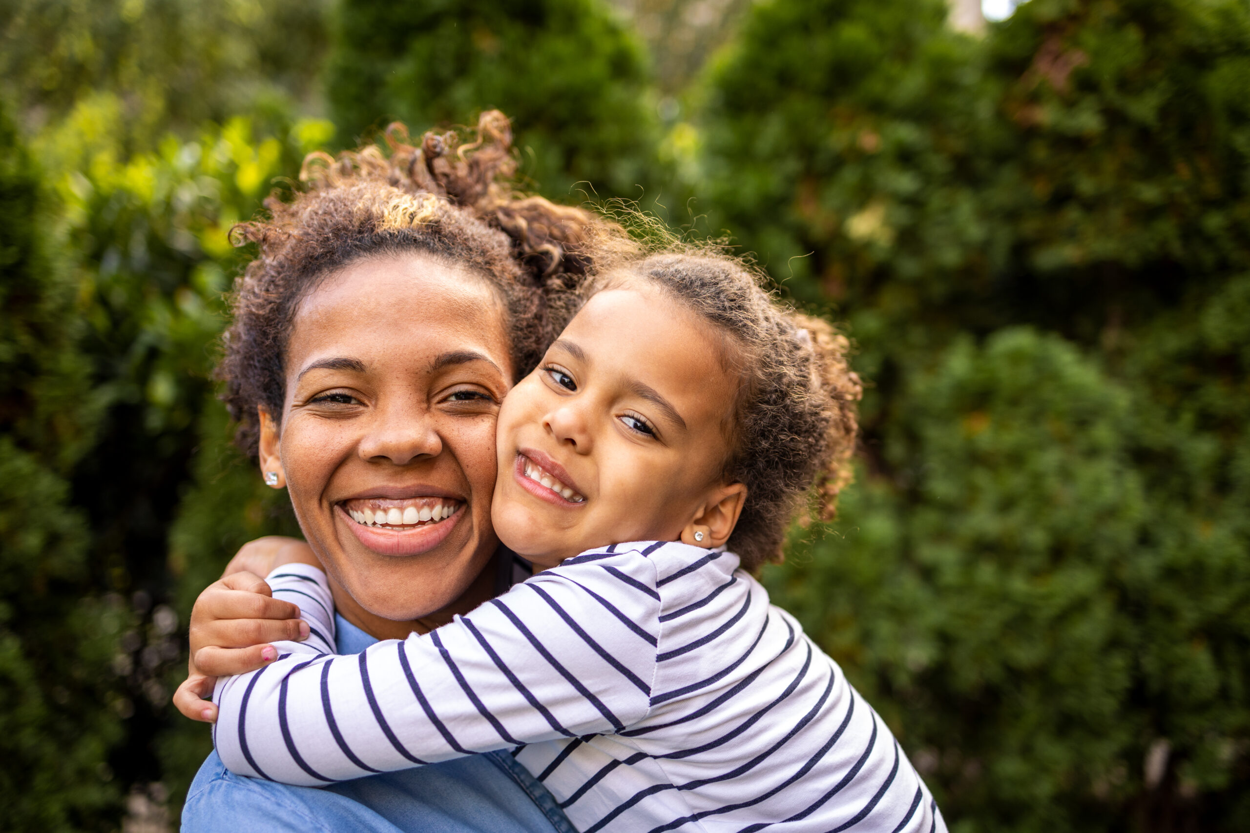 A mom holds her young daughter outside on a sunny day, both smiling and happy.