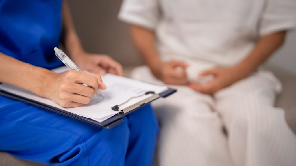 A nurse and patient sit together. The nurse takes notes on a clipboard while the patient talks.