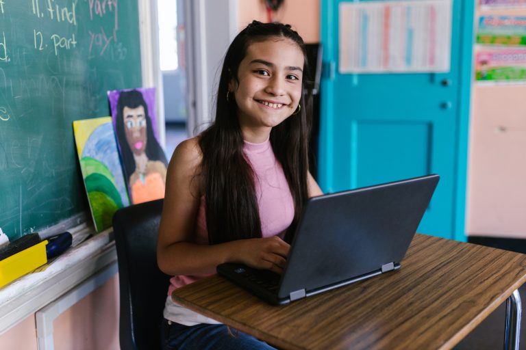 A young person smiles over their laptop screen in a classroom